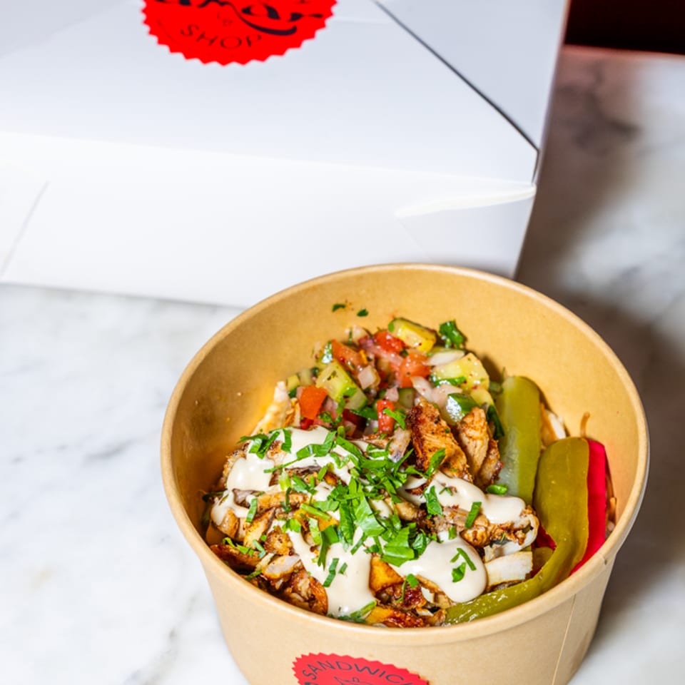 Shawarma bowl with chicken, vegetables, sauce, and pickles, next to a white takeout box.
