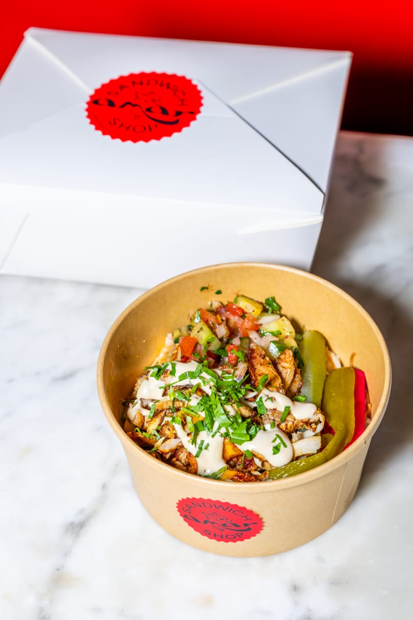 Shawarma bowl with chicken, vegetables, sauce, and pickles, next to a white takeout box.