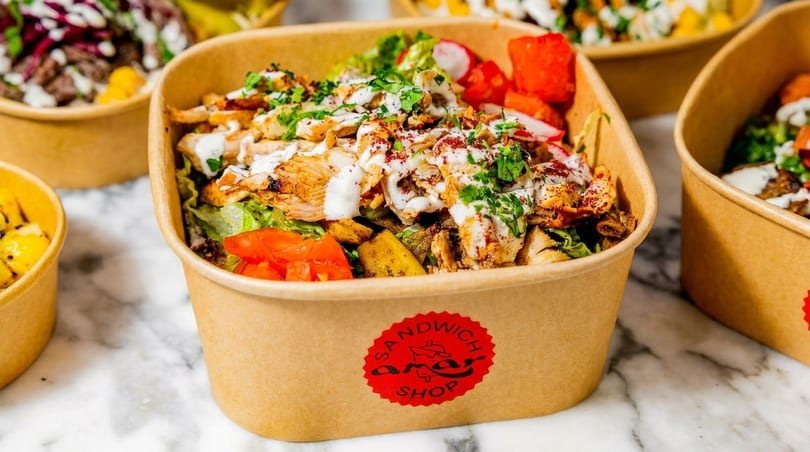 A brown takeaway box filled with chicken shawarma, lettuce, tomatoes, and white sauce, on a marble surface.