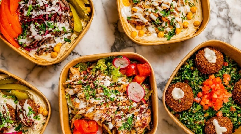 Four bowls of Middle Eastern food, including shawarma, falafel, and salads, on a marble surface.