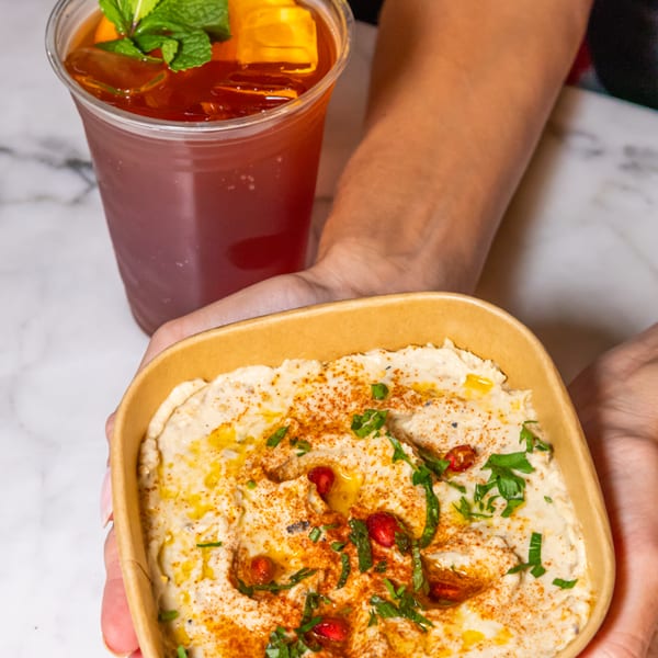 Hands holding a bowl of hummus with paprika, pomegranate, and parsley, next to an iced tea.