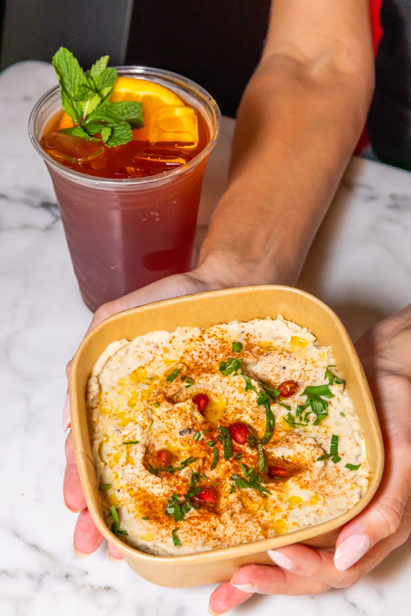 Hands holding a bowl of hummus with paprika, pomegranate, and parsley, next to an iced tea.
