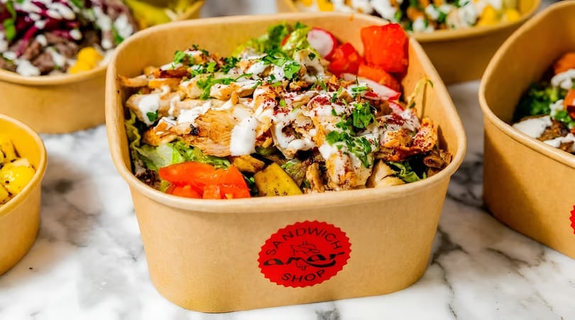 A close-up of a chicken bowl from Sandwich Shop, topped with sauce, herbs, and vegetables.