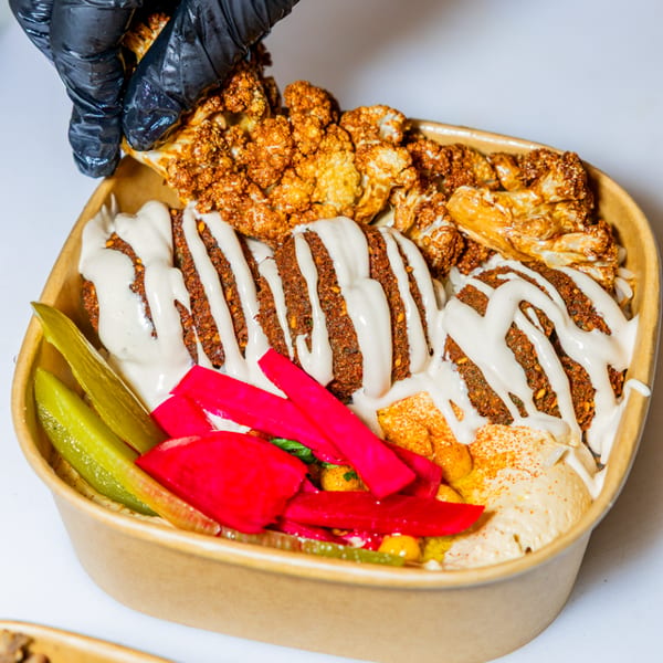 A gloved hand adds roasted cauliflower to a falafel bowl with hummus, pickles, and tahini.