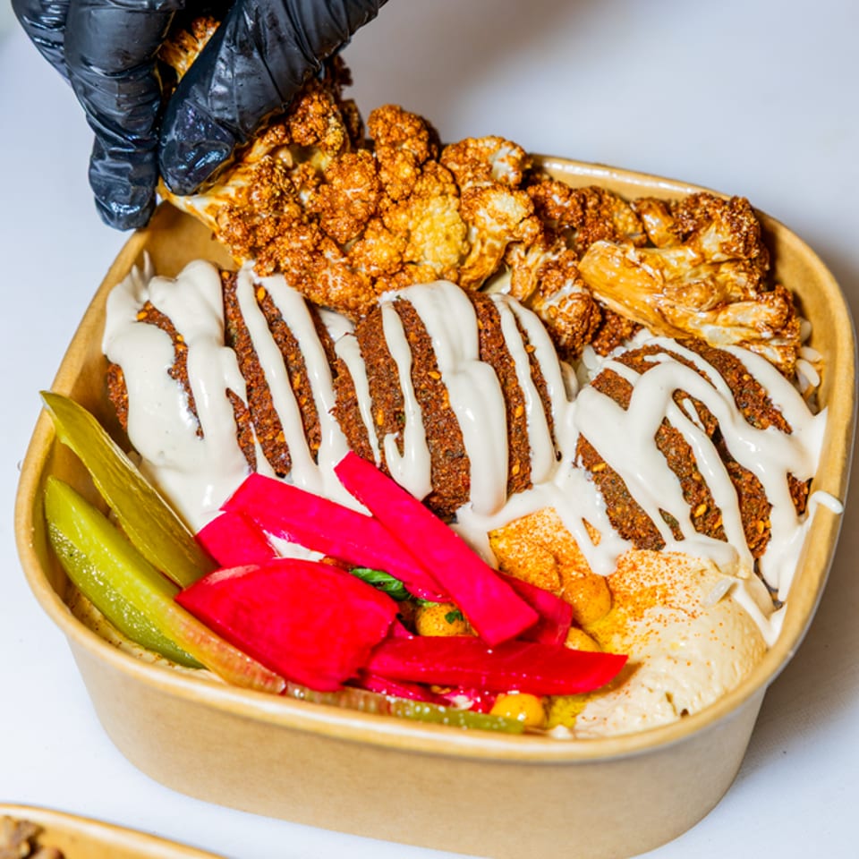 A gloved hand adds roasted cauliflower to a falafel bowl with hummus, pickles, and tahini.