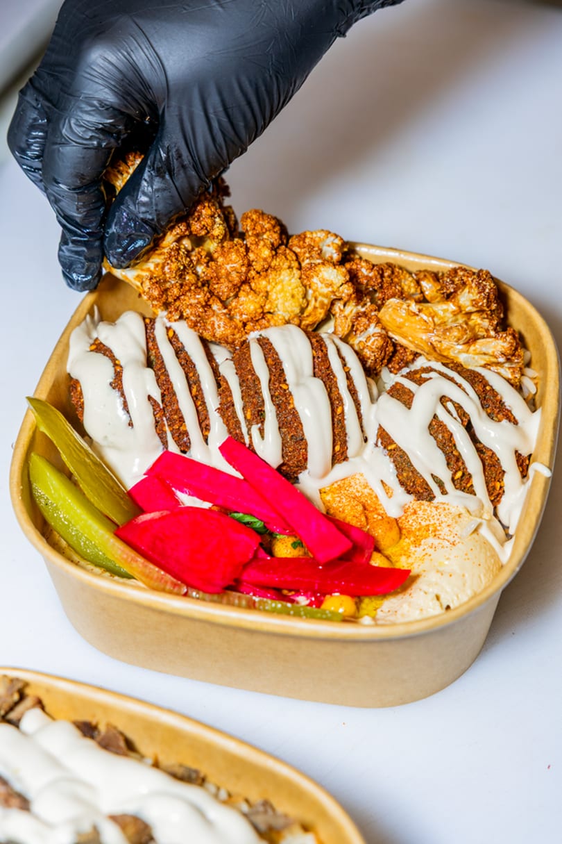 A gloved hand adds roasted cauliflower to a falafel bowl with hummus, pickles, and tahini.