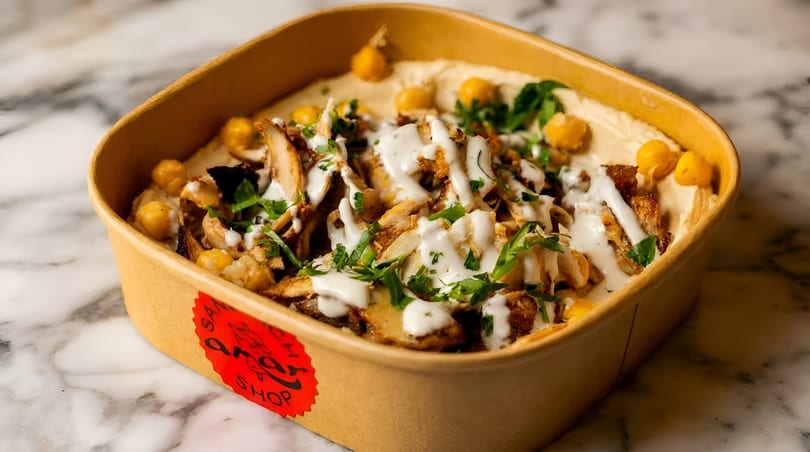 Shawarma hummus bowl with chicken, chickpeas, parsley, and white sauce in a brown container.